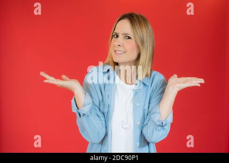 Junge Frau, die über rot isoliert Hintergrund ahnungslos und verwirrt Ausdruck mit offenen Armen. Zweifelskonzept. Stockfoto