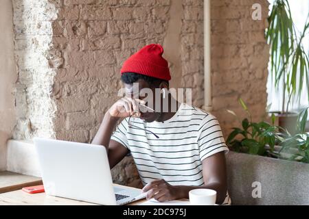 Millennial Afro-American Mann in roten Hut tragen drahtlose Kopfhörer, hält Brille, reiben seine Augen, fühlt sich müde nach der Arbeit am Laptop im Café, taki Stockfoto