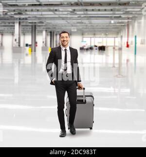 Porträt eines lächelnden jungen Mannes in einem Schwarzer Anzug, der in einer leeren Flughafenhalle mit einem läuft Koffer Stockfoto