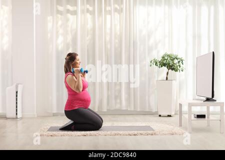 Schwangere Frau kniend und Training mit Hanteln vor fernseher zu Hause Stockfoto