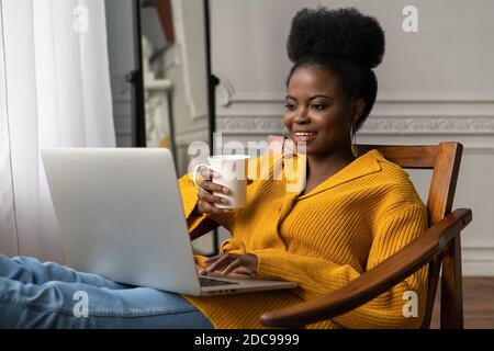 Happy Afro-amerikanische Millennial Frau mit Afro Frisur tragen gelbe Strickjacke ruhen, sitzen auf einem Stuhl, beobachten Webinar, arbeiten online auf Laptop, t Stockfoto