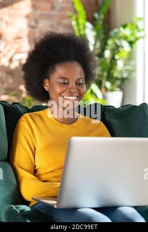 Lächelnde afroamerikanische Biracial junge Frau mit afro Frisur in gelben Pullover sitzen in grünen Stuhl, Remote-Arbeit auf Laptop, Chat und Talkin Stockfoto