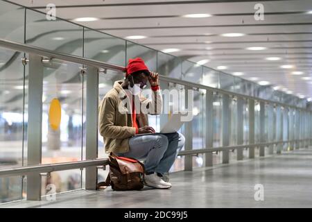 Stilvolle Black Traveler Millennial Mann tragen Gesicht Schutzmaske, sitzen in leeren Flughafen-Terminal, funktioniert Remote auf Laptop, während auf einen Flug warten Stockfoto