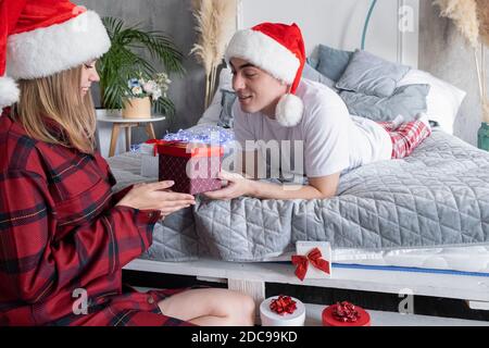 Weihnachten, Urlaub, Winter und Menschen Konzept. Glückliches Paar mit weihnachtsgeschenk zu Hause tragen weihnachtsmützen. Junger lächelnder Mann, der auf dem Bett liegt Stockfoto
