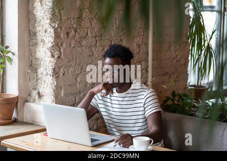 Fokussierte Millennial Black man Student in T-Shirt Stripped Brille tragen, beobachten pädagogische Webinar auf Laptop, Remote-Online-Arbeit im Café, Studium, Stockfoto