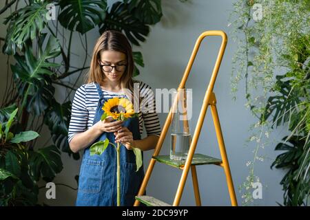 Floristin Frau umgeben von tropischen Pflanzen halten Sonnenblume, in der Nähe der orangefarbenen Trittleiter in ihrem Blumengeschäft stehen. Stockfoto
