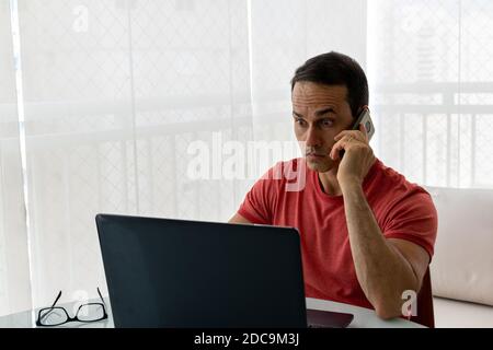 Reifer Mann mit Handy in der Hand, Blick auf Laptop mit weiten Augen. Stockfoto