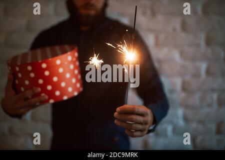 Nahaufnahme eines Neujahrssprinklers in den Händen eines jungen Mannes, der in zwangloser Kleidung eine rote Geschenkbox hält. Frohe Feiertage Stockfoto