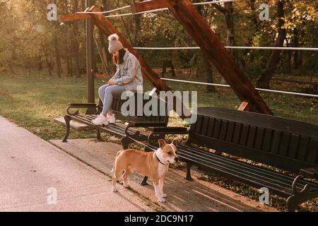 Ein süßes junges Teenager-Mädchen mit Hut sitzt auf einer Parkbank und tippt eine Nachricht am Telefon, und neben ihr ist ein brauner Hund Stockfoto