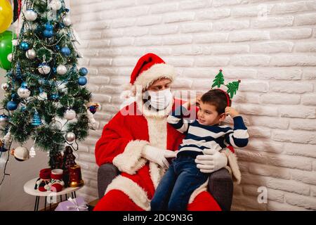 Ein fröhlicher 5-jähriger Junge mit einer Requisite auf dem Kopf Sitzen in Santa's Schoß mit einer medizinischen Gesichtsmaske.Überraschung für Ein Kind während des COVID-19 Coronavirus Stockfoto