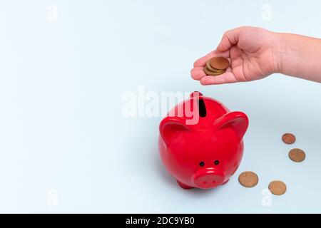 Kinderhand hält mehrere Münzen über einem roten Sparschwein neben ihnen auf einem blauen Hintergrund liegen, Draufsicht, Kopierraum. Sparkonzept Stockfoto