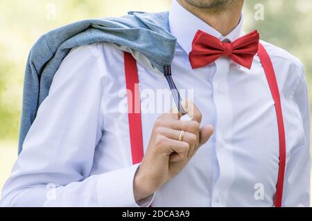 Der Bräutigam im weißen Hemd mit roter Schleife Krawatte und in Hosenträger hält eine Jacke Stockfoto