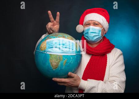 Zeigt das Zeichen eines Shootings. Europäischer Mann in einer medizinischen Maske und einer roten Weihnachtsmütze, die einen Globus in einer medizinischen Maske auf schwarzem Hintergrund hält. Pandemisches Coronavirus, Quarantäne, 2020, 2021. . Hochwertige Fotos Stockfoto