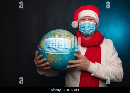 Europäischer Mann in einer medizinischen Maske und einer roten Weihnachtsmütze, die einen Globus in einer medizinischen Maske auf schwarzem Hintergrund hält. Pandemisches Coronavirus, Quarantäne, 2020, 2021. Feiern Sie das neue Jahr, zu Hause bleiben, . Hochwertige Fotos Stockfoto