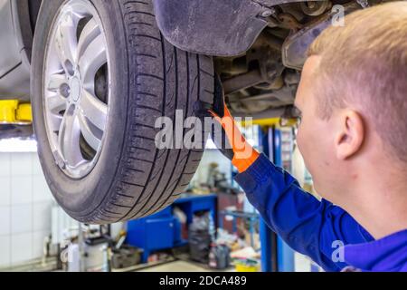 Nahaufnahme des Mechanikers, der die Oberfläche des Autoreifens sehr aufmerksam überprüft. Mechaniker mit blauem Overall und orangefarbenen Handschuhen. Auto ist auf einem hydraulischen lif Stockfoto