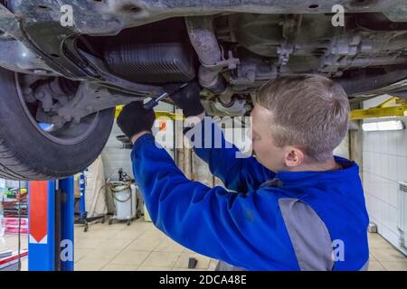 Mechaniker, der unter einem Auto in der Garage arbeitet. Techniker mit blauem Overall und Schraubenschlüssel. Das Auto befindet sich auf einem hydraulischen Aufzug. Stockfoto