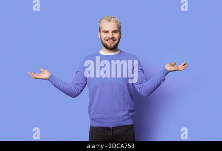 Monochromes Foto eines bärtigen Mannes mit blondem Haar beim Vergleich Zwei Dinge auf seinen Handflächen lächeln auf einem blauen Studio Wand Stockfoto