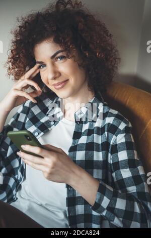 Die lockige Frau liegt im Sessel und lächelt an Kamera beim Chatten auf einem Telefon Stockfoto