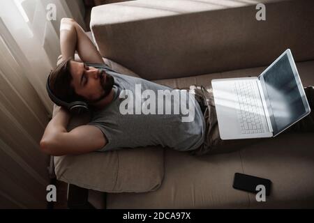 Mann auf dem Sofa mit Kopfhörer und Notebook zu Hause. Junge Hipster tragen Kopfhörer Studie online beobachten Webinar Podcast auf Laptop hören Lernen edu Stockfoto