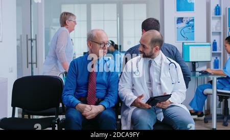 Arzt erklärt dem älteren Mann im Wartebereich des Krankenhauses die Behandlung. Frau mit Gehrahmen bei der medizinischen Untersuchung ankommen. Stockfoto