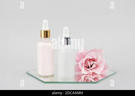 Zwei Glasflaschen mit Serum, ätherischem Öl oder anderem Kosmetikprodukt und schönen rosa Blüten auf Spiegel auf grauem Hintergrund. Natürliches Organisches Spa Co Stockfoto