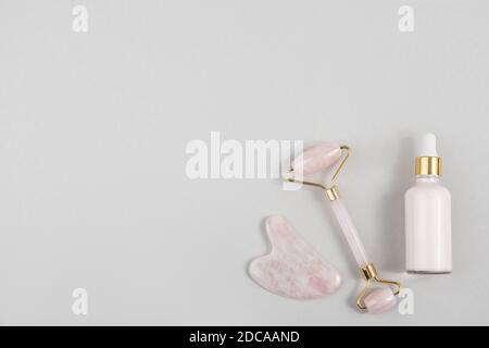 Crystal Rose Quarz Gesichtswalze, Massage-Tool Gua Sha und Anti-Aging-Kollagen, Serum in Glasflasche auf grauem Hintergrund. Gesichtsmassage für natürliche Stockfoto