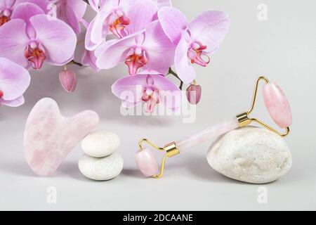 Crystal Rose Quarz Gesichtswalze und Massage-Tool Jade Gua Sha auf Steinen und natürliche Orchidee Blume auf grauem Hintergrund. Gesichtsbehandlung Anti-Age-Massage für na Stockfoto