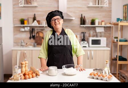 Alte Dame mit Kochschürze und bonete in der Küche mit Mehl auf dem Tisch. Rentner älterer Bäcker in der Küche Uniform Vorbereitung Gebäck Zutaten auf Holztisch bereit, hausgemachte leckeres Brot kochen. Stockfoto