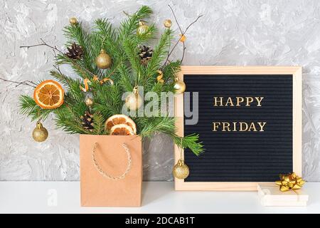 Happy Friday Text auf schwarzem Briefbrett und festlicher Blumenstrauß aus Tannenzweigen mit weihnachtsdekor in Bastelpackung und Geschenkbox auf Tisch. Vorlage für Stockfoto