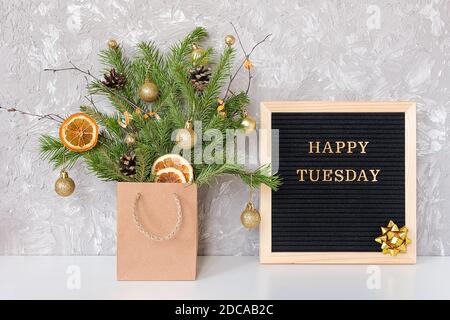 Happy Tuesday Text auf schwarzem Briefbrett und festlicher Blumenstrauß von Tannenzweigen mit weihnachtsdekor in Bastelpackung auf Tisch. Vorlage für Postkarte, gr Stockfoto
