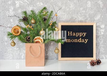Happy Monday Text auf schwarzem Briefbrett und festlicher Blumenstrauß aus Tannenzweigen mit weihnachtsdekor in Bastelpackung auf Tisch. Vorlage für Postkarte, gre Stockfoto