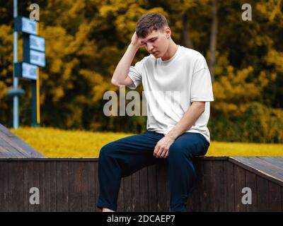 Weißes T-Shirt Mockup auf jungen Kerl, der seine Haare gerade, sitzt vor verschwommenem Straßenhintergrund, Vorderansicht. Leere stilvolle Kleidung Vorlage für de Stockfoto