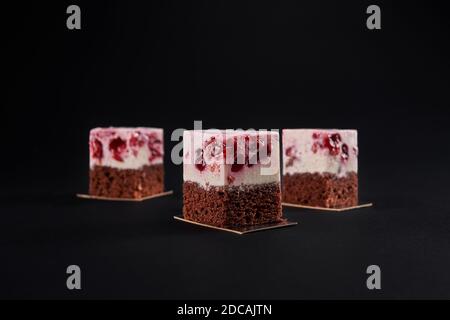 Nahaufnahme von drei leckeren frischen Kuchenscheiben mit braunem Keks und Mousse rosa Schicht mit rotem Gelee und Kirschen. Leckere süße Dessert isoliert auf schwarzem Hintergrund im Restaurant. Stockfoto