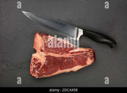 Nahaufnahme eines marmorierten rohen Porterhouse T-Bone Beef Steak mit Rippenknochen auf schwarzem Schiefer Schneidebrett mit Messer, erhöhte Draufsicht, direkt darüber Stockfoto