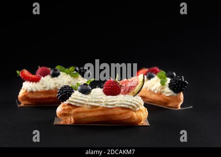 Gebackene hausgemachte Ecolirs gefüllt mit Sahne, weißen Belag und Scheiben von Feigen, Brombeeren, Himbeeren und Heidelbeeren. Seitenansicht von frischem Dessert in Reihe isoliert auf schwarzem Hintergrund. Stockfoto