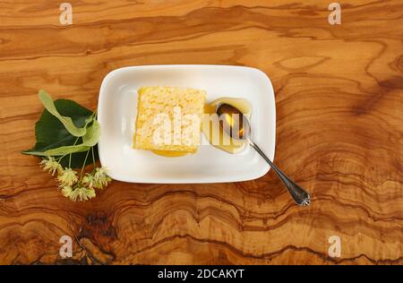 Nahaufnahme frischer Akazienhonig mit Honigwabe, Löffel und Lindenblüten auf weißem Teller über Holztisch, erhöhte Draufsicht, direkt darüber Stockfoto