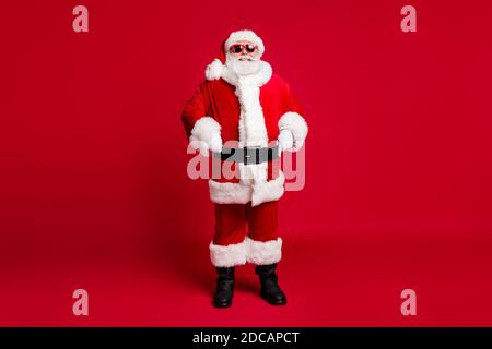 In voller Länge Foto von Rentner alten Mann grauen Bart selbstsicher Hände Hüften weihnachtsbaum Ereignis tragen santa Kostüm Handschuhe Mantel Kopfbedeckung Sonnenschutzscheibe des Bandes Stockfoto