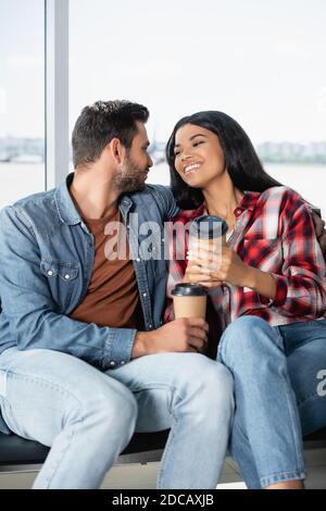 Glückliches multikulturelles Paar, das Kaffee hält, um zu gehen, während es innen sitzt Abflughalle des Flughafens Stockfoto