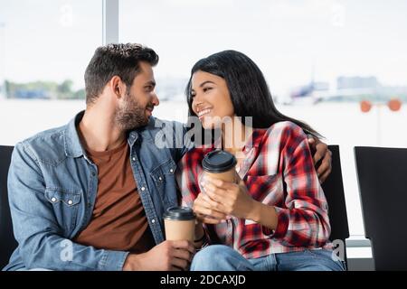 Lächelndes multikulturelles Paar, das Kaffee hält, um zu gehen, während es innen sitzt Abflughalle des Flughafens Stockfoto