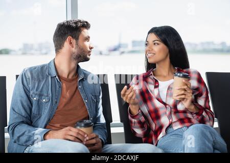 Fröhliches multikulturelles Paar, das Kaffee hält, um zu gehen, während es innen sitzt Abflughalle des Flughafens Stockfoto