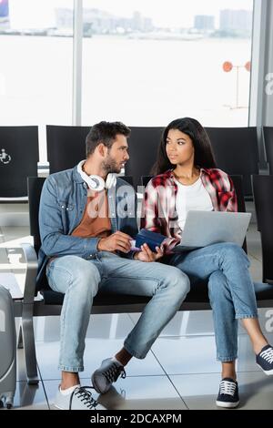 Bärtiger Mann mit Kopfhörern, der Pass hält und afrikaner anschaut amerikanische Frau mit Laptop am Flughafen Stockfoto