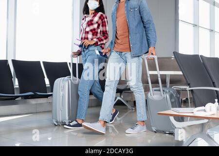 Beschnittene Ansicht der afroamerikanischen Frau in der medizinischen Maske halten Pass mit Bordkarte beim Spaziergang mit Mann im Flughafen Stockfoto