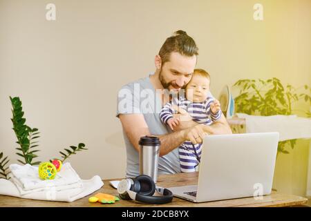 Kaukasischer Mann, der zu Hause mit einem Laptop und einem Baby an den Händen arbeitet. Bleib zu Hause Konzept. Freiberufliches und selbstisolierende Konzept. Stockfoto