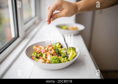 Hand hinzufügen eine Prise Salz zu einem Brokkoli und Hühnersalat neben einem Fenster Stockfoto