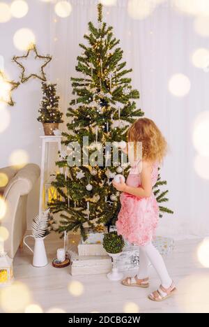 Unerkennbar 6 Jahre altes Mädchen in rosa Kleid, hängende Weihnachtskugel Ornamente auf Weihnachtsbaum. Weißes, minimalistisches Wohnzimmer. Stockfoto