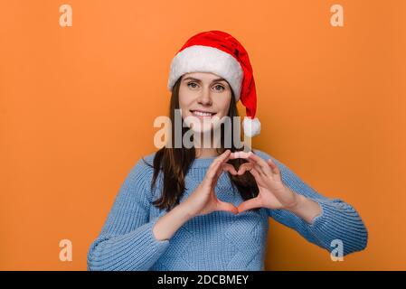 Hübsche junge Frau im weihnachtlichen roten Hut, macht Herzgeste, zeigt ihre wahren Gefühle, hat fröhlichen Ausdruck, trägt Winterpullover Stockfoto