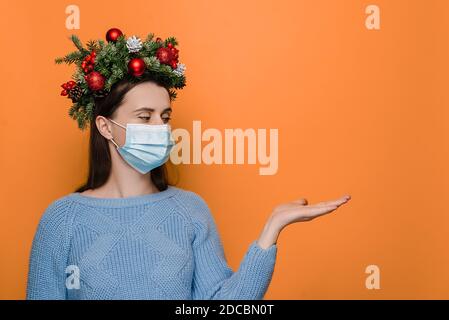 Junge kaukasische Frau in medizinischen Gesichtsmaske, hält die Hand angehoben, in Winterpullover und handgefertigten Kranz gekleidet, gibt vor, etwas zu halten Stockfoto