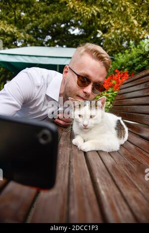 Ein Mann macht ein Selfie mit einer streunenden Katze sitzend Auf einer Bank Stockfoto