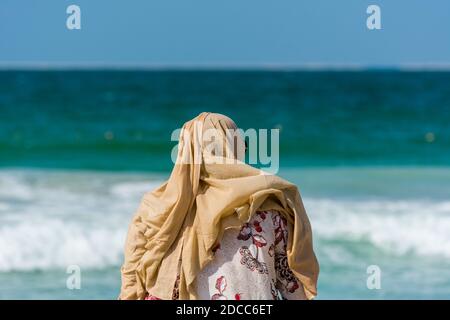 Eine islamische Frau trägt Hijab und beobachtet die Wellen, Wasser spritzt Spülung Strand am Jumeirah Strand am Persischen Golf in Dubai, Vereinigte Arabische Emirate. Stockfoto