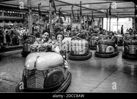 Ein Teenager-Paar lächelt, als sie ihr Dodgem-Auto vor den folgenden Autos auf einem Messegelände in den 1950er Jahren fahren Stockfoto